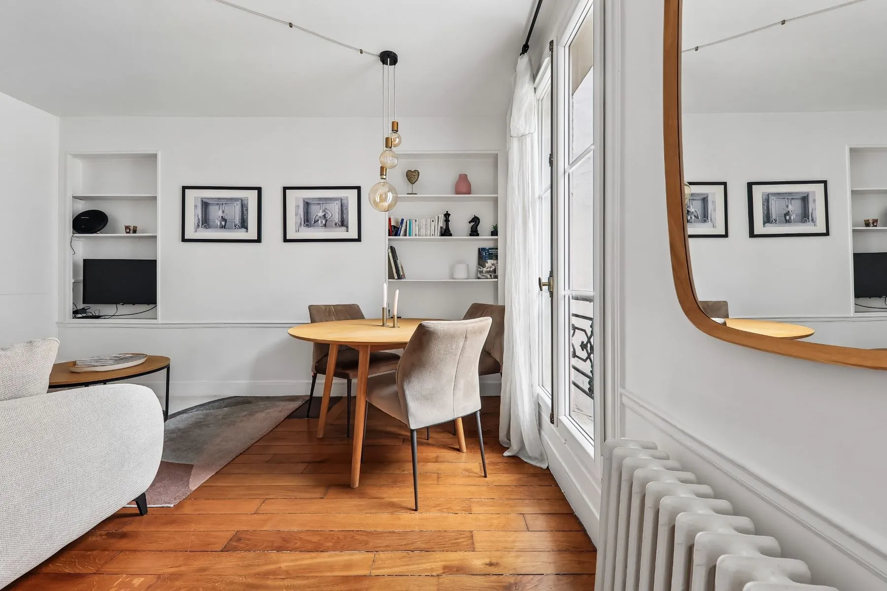 Rounded dining table beside tall windows, timber-framed mirror and cast-iron radiator