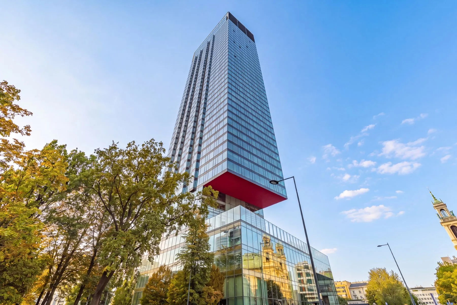 Glass exterior of Cosmopolitan Tower rising above trees and neighbouring buildings in Warsaw