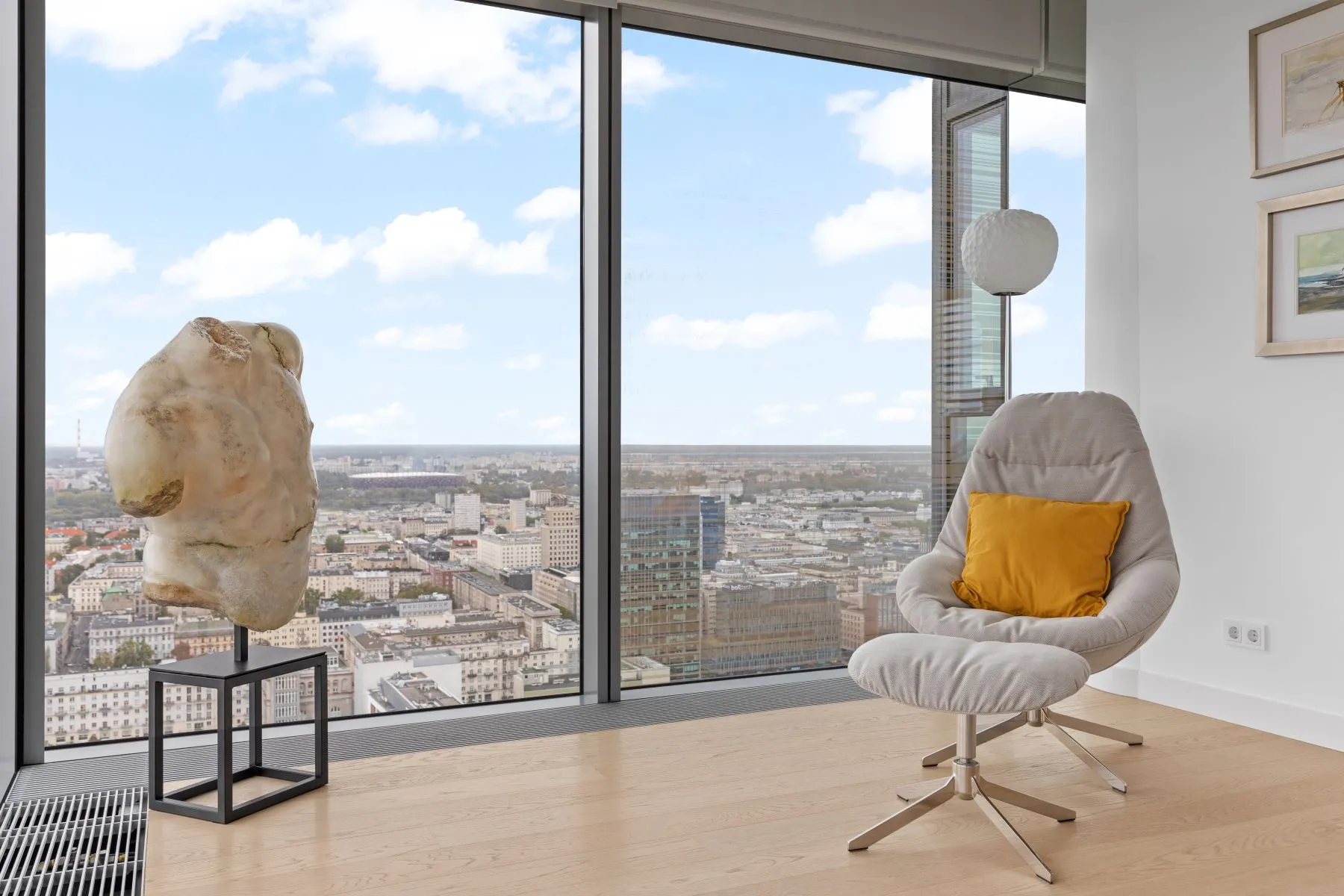 Grey lounge chair with yellow cushion beside a stone sculpture and wide city-facing windows