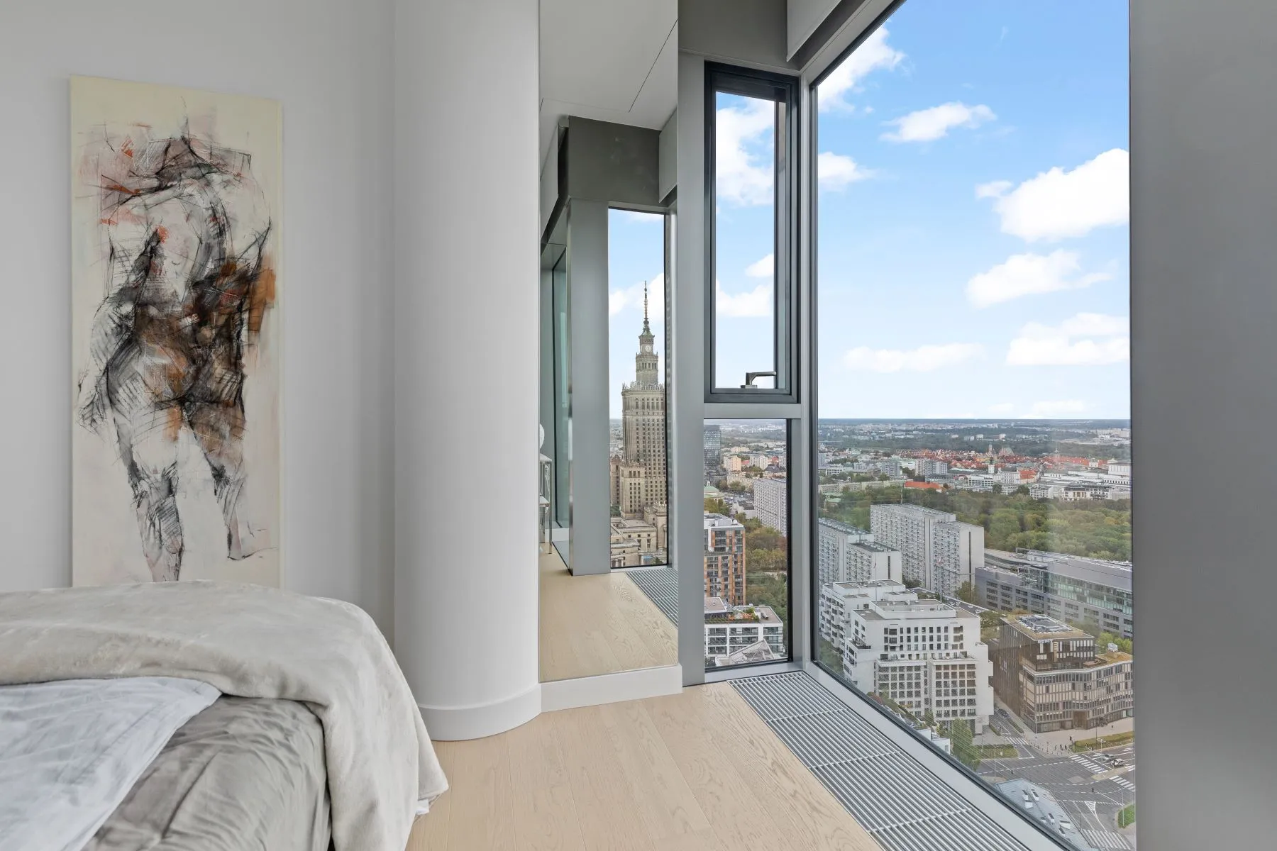 Bedroom corner with artwork, mirrored storage and a tall window overlooking central Warsaw
